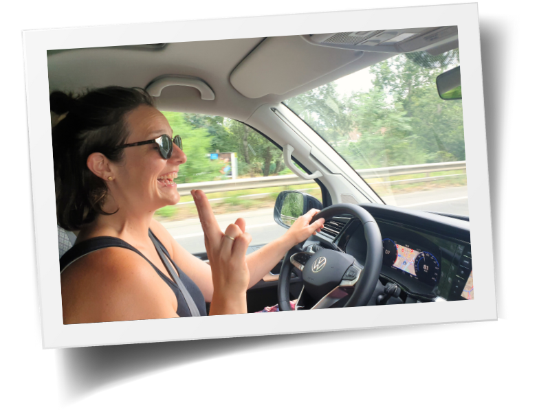Polaroidfoto aus dem Inneren von einem VW-Bus während der Fahrt, Beifahrerperspektive. Eine Frau mit mittellangem, braunem Haar steuert das Fahrzeug. Sie trägt eine Sonnenbrille, lacht und trägt ein graues Top. Mit der linken Hand steuert sie das Fahrzeug. Die rechte Hand zeigt ein Peace-Zeichen.