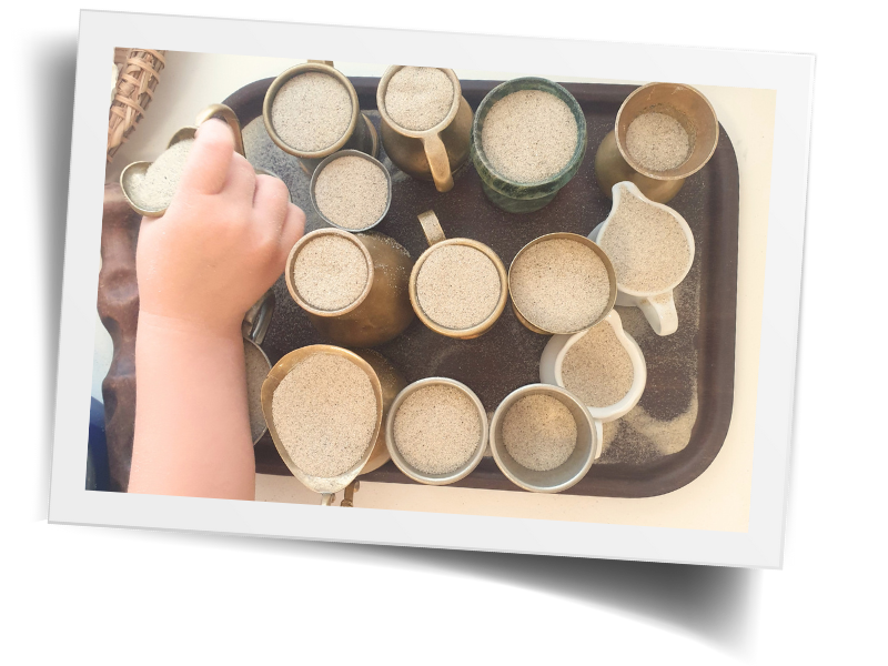 Foto mit weißem Rand, aus Vogelperspektive: Kinderarm, der nach einer von mehreren, mit Sand gefüllten, Kannen und Vasen auf einem silbernen Tablett greift. 