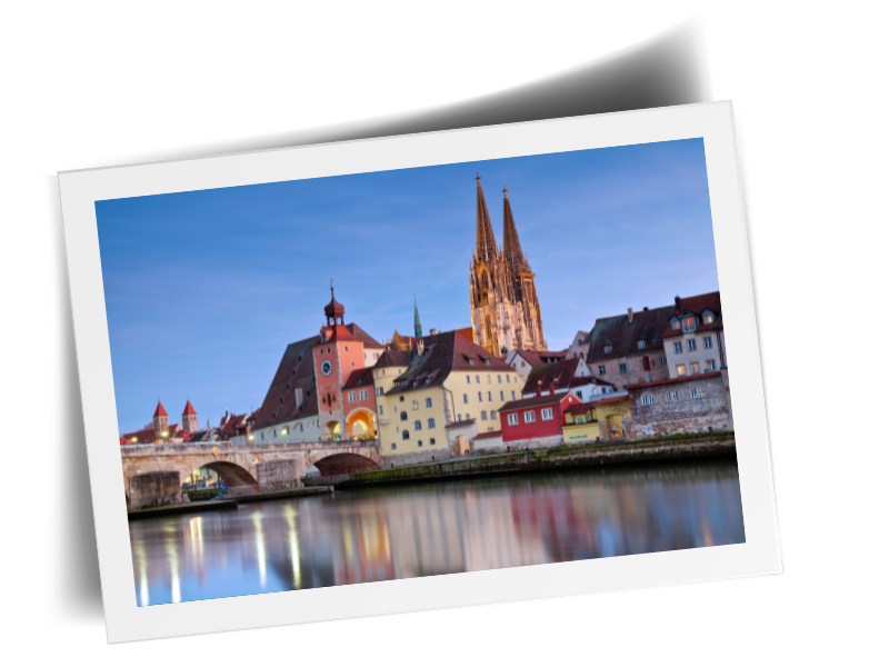 Aufnahme der Regensburger Altstadt bei Dämmerung. Perspektive von der Gegenüberliegenden Seite des Flusses. Zu sehen sind bunte Häuser, eine Brücke und eine Kirche im Hintergrund der Regensburger Dom.